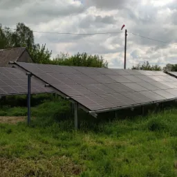 Panele fotowoltaiczne zamontowane na konstrukcji w Mławie, widoczne na tle wiejskiego krajobrazu z budynkiem gospodarczym i drzewami.