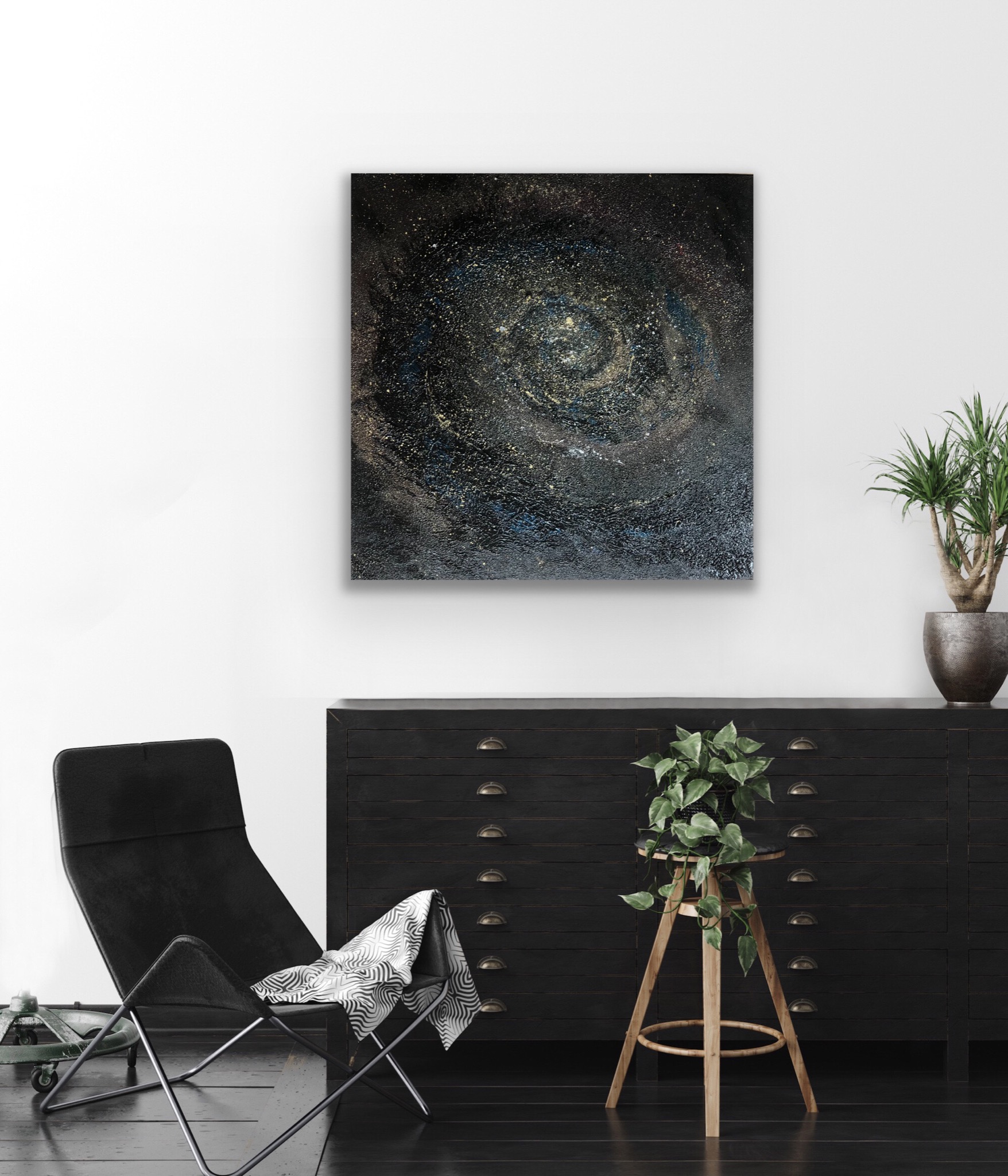 Abstrakcyjne malowidło w odcieniach czerni, złota i błękitu, przypominające galaktykę, zawieszone na białej ścianie nad czarną komodą z szufladami, obok czarnego krzesła i rośliny doniczkowej.