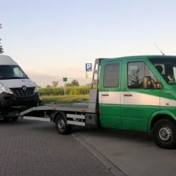 Zielona laweta z białym pasem, na stacji benzynowej, przewozi biały samoch&oacute;d dostawczy DPD.