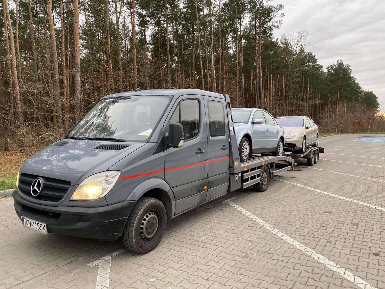 Szara laweta Mercedes-Benz z dwoma osobowymi samochodami na platformie, zaparkowana na parkingu z kostki brukowej w pobliżu lasu.