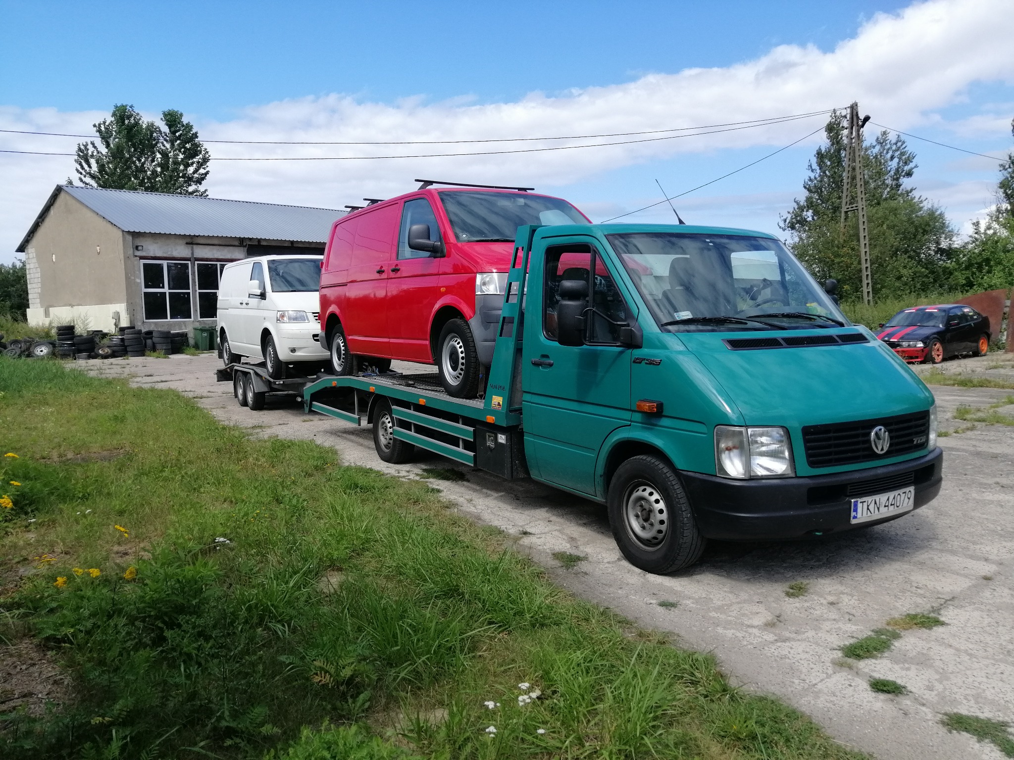 Zielona laweta z dwoma samochodami dostawczymi: czerwonym na platformie i białym na przyczepie, na tle budynku warsztatowego i zieleni.