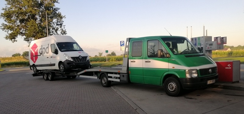 Zielona laweta z białym pasem, na stacji benzynowej, przewozi biały samochód dostawczy DPD.