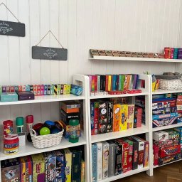 Centrum Edukacji i Turystyki EWART - Półki wypełnione grami edukacyjnymi i językowymi, w tym gry planszowe i karciane, z tabliczkami 'Welcome' na ścianie w tle.