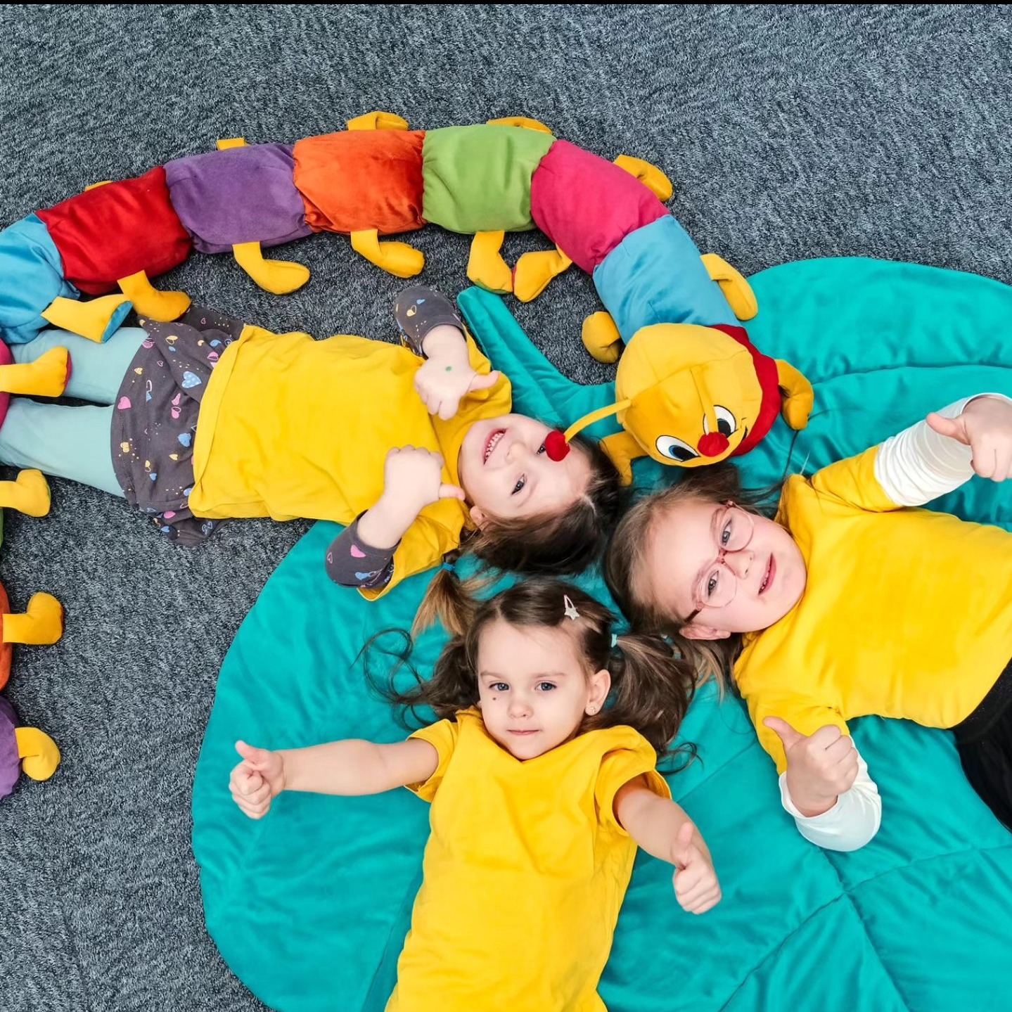 Trzy uśmiechnięte dziewczynki w żółtych koszulkach leżą na turkusowej poduszce z pluszową, kolorową gąsienicą, pokazując kciuki w górę, na tle szarego dywanu.