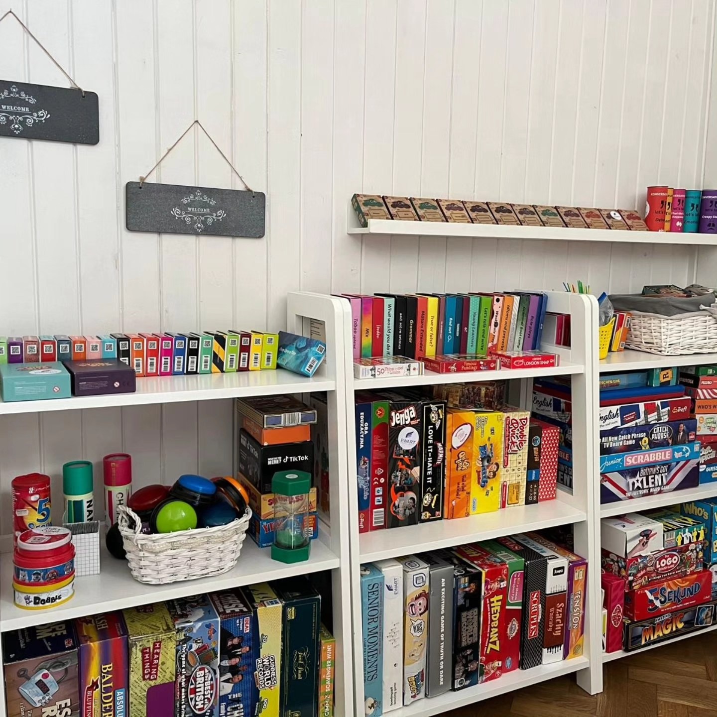 Półki wypełnione grami edukacyjnymi i językowymi, w tym gry planszowe i karciane, z tabliczkami 'Welcome' na ścianie w tle.