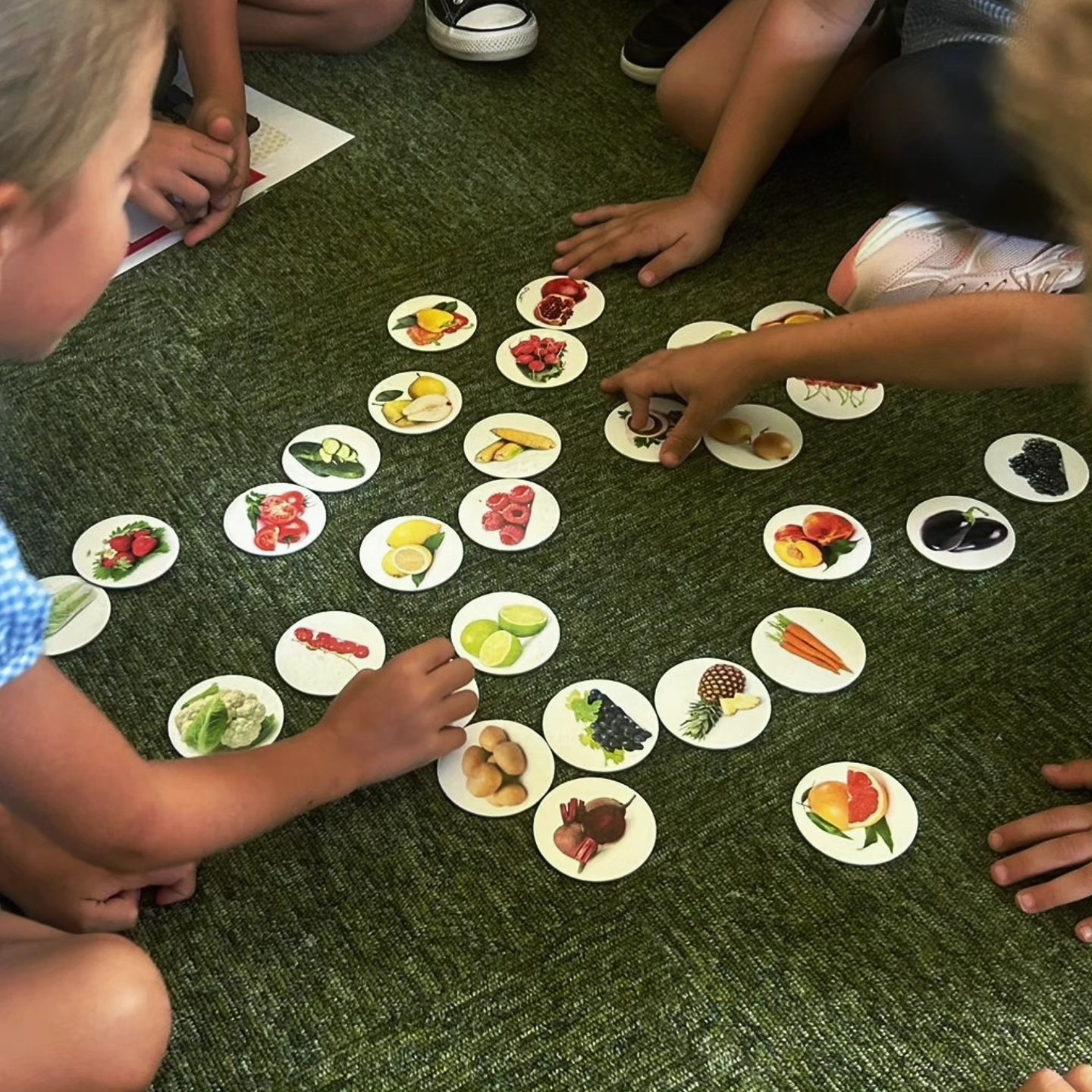 Dzieci grają w grę edukacyjną z kartami przedstawiającymi owoce i warzywa, rozłożonymi na zielonym dywanie.