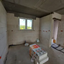 Wnętrze w stanie surowym z widocznymi bloczkami betonowymi, oknem z widokiem na zieleń, puszkami elektrycznymi w ścianach i przewodami na podłodze; paczka bloczków z widocznym logo producenta...