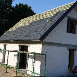 Wznoszony dom jednorodzinny z białych bloczków, częściowo pokryty ciemną dachówką, widoczne rusztowania i składowane bloczki.