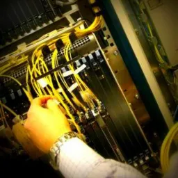 Instalacja światłowodów w serwerowni: ręka technika podłącza żółte kable światłowodowe do panelu krosowniczego w szafie rack, widoczny zegarek na nadgarstku.