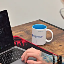 Osoba z tatuażem na ręce pracuje na laptopie MacBook Pro; na ekranie widoczne logo firmy Binarest, a obok stoi kubek z logo tej samej firmy, całość na drewnianym biurku z podkładką pod mysz...