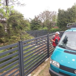 Ddshops Jarosław Dębski - Nowoczesne, poziome, szare ogrodzenie panelowe z metalu, widoczne z boku; obok zaparkowany turkusowy van z bagażnikiem dachowym, a mężczyzna w czerwonej kamizelce czyści przednią szybę.