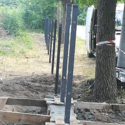 Ddshops Jarosław Dębski - Montaż czarnych słupków ogrodzeniowych w betonowej podstawie, widoczne drewniane szalunki i biały samochód dostawczy w tle, teren budowy.