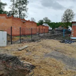 Budowa domu jednorodzinnego w trakcie realizacji: widoczne mury z czerwonej cegły, ogrodzenie z czarnej siatki oraz skrzynka elektryczna przyłączeniowa na placu budowy, w tle domy mieszkalne.