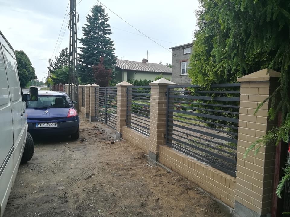 Nowoczesne ogrodzenie z ceglanymi słupkami i poziomymi metalowymi przęsłami, widoczne z perspektywy ulicy, z zaparkowanym samochodem i bujną zielenią w tle.