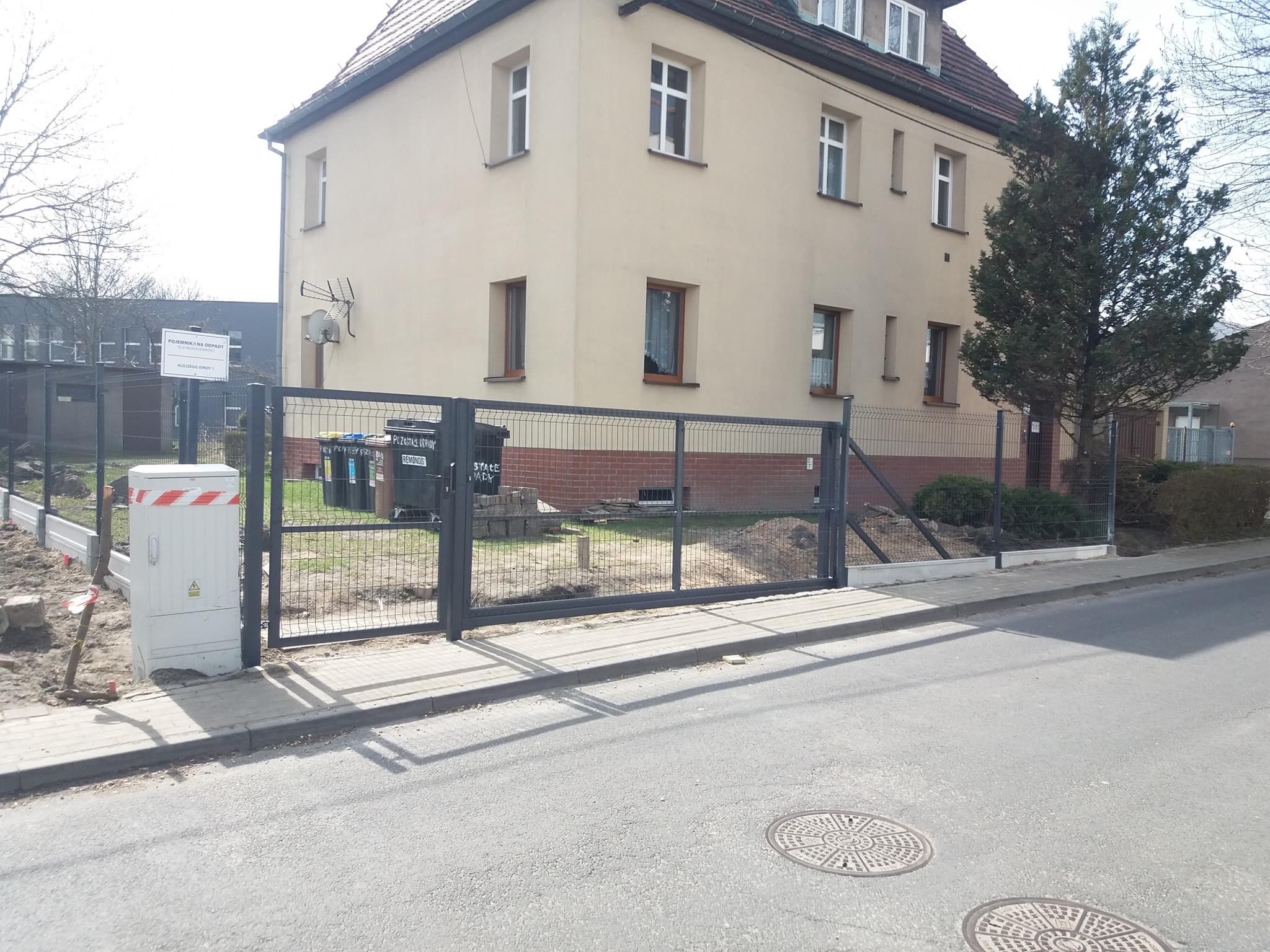 Szara metalowa brama panelowa przed budynkiem z beżową elewacją, widoczne ślady prac ziemnych i przygotowania terenu, skrzynka elektryczna zabezpieczona taśmą ostrzegawczą.