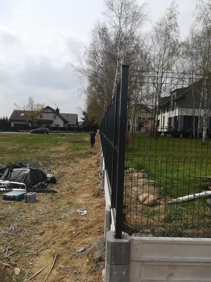 Montaż ogrodzenia panelowego z podmurówką betonową, widoczne narzędzia i materiały budowlane na placu budowy, w tle domy jednorodzinne i drzewa.