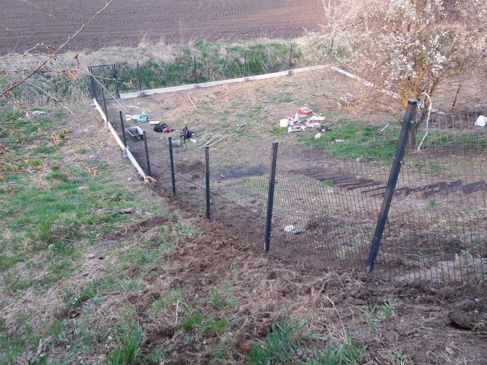 Montaż ogrodzenia panelowego na nierównym terenie, widoczne słupki i fragment siatki, w tle narzędzia i materiały budowlane, drzewo kwitnące w tle.