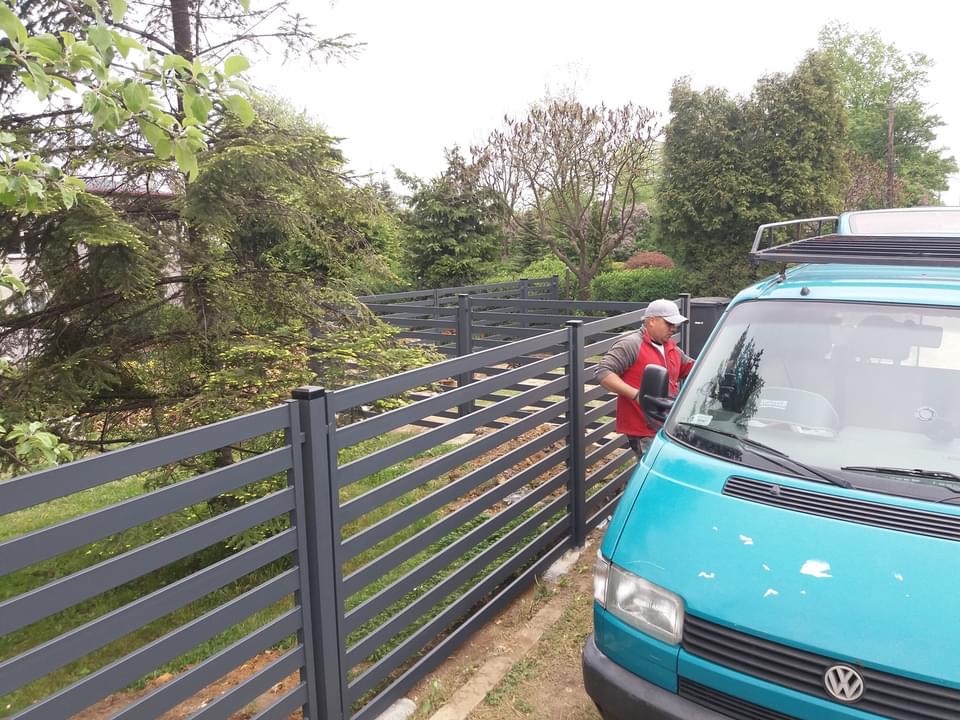 Nowoczesne, poziome, szare ogrodzenie panelowe z metalu, widoczne z boku; obok zaparkowany turkusowy van z bagażnikiem dachowym, a mężczyzna w czerwonej kamizelce czyści przednią szybę.