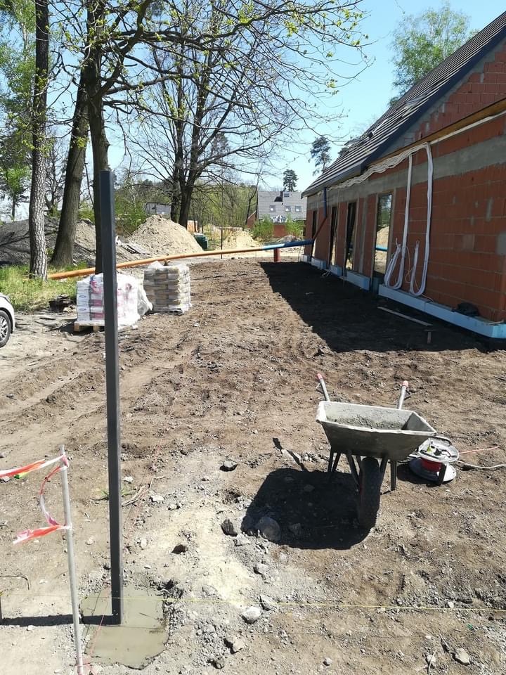 Budowa domu jednorodzinnego w stanie surowym otwartym: widoczne ściany z czerwonej cegły, brak okien, wystające przewody elektryczne, teren przygotowany pod elewację, stos kostki brukowej i taczka...