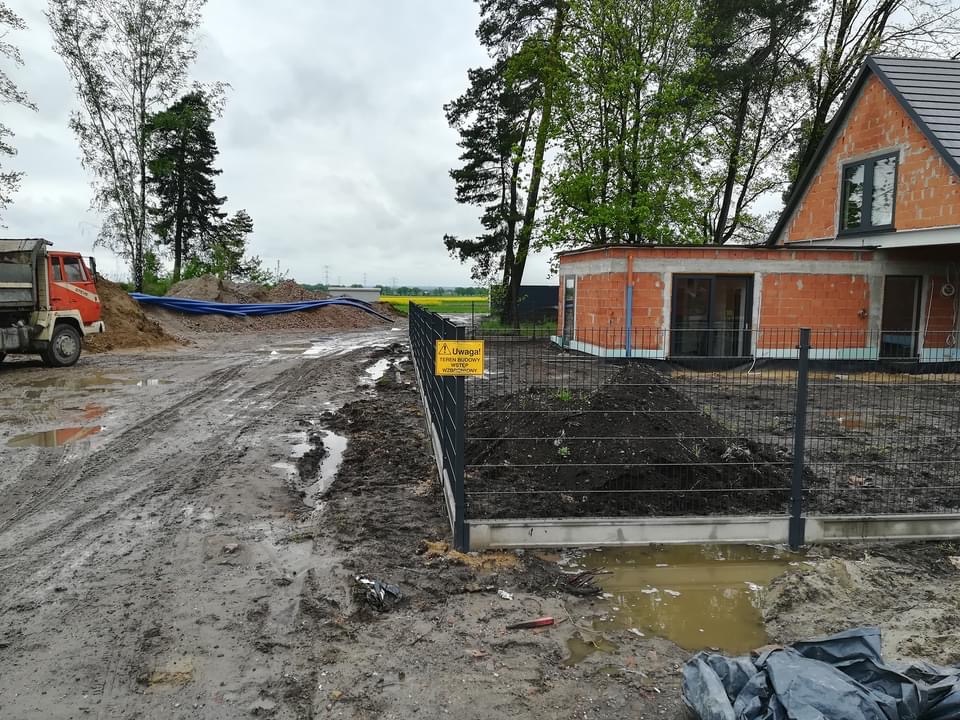 Budowa domu w trakcie realizacji: widok na surową elewację z czerwonej cegły, ogrodzenie panelowe z tabliczką ostrzegawczą, błotnista droga dojazdowa i wywrotka w tle.