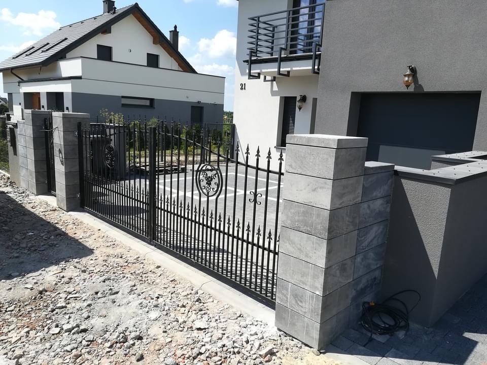 Czarna, metalowa brama wjazdowa z ozdobnymi elementami, osadzona na szarych, betonowych słupkach, prowadząca na posesję z nowoczesnym domem dwukondygnacyjnym.