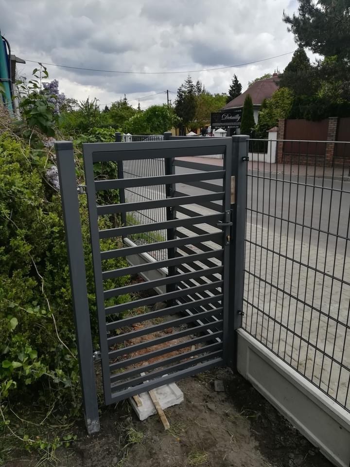 Nowoczesna, szara furtka z poziomymi szczeblami, zamontowana przy ogrodzeniu panelowym, na nierównym terenie z kostką betonową i resztkami gruzu. W tle widoczna zieleń ogrodu i budynek.