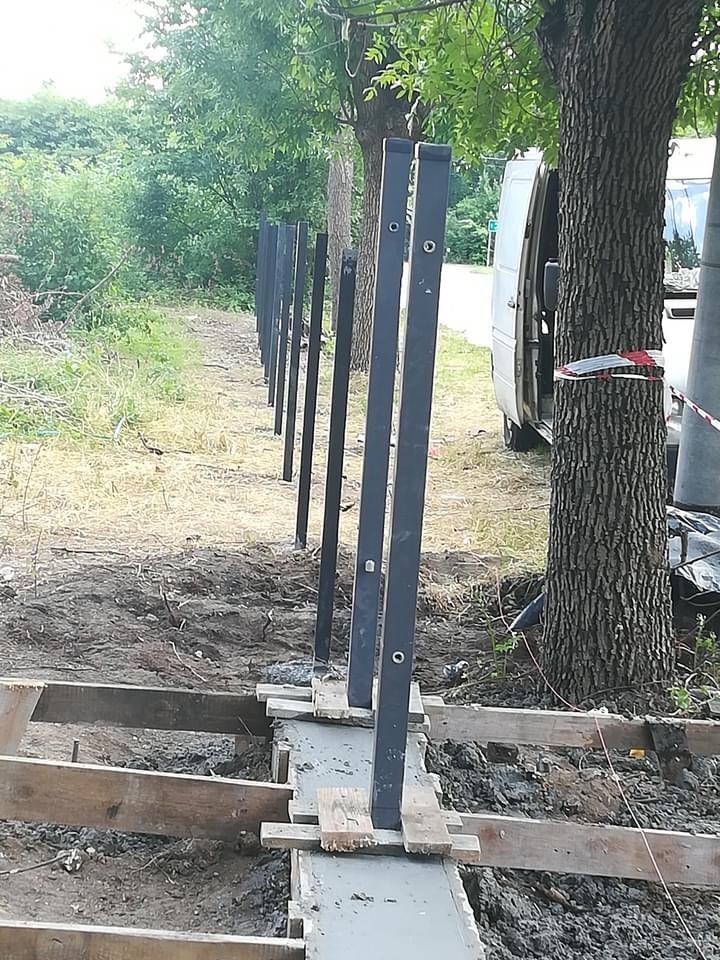 Montaż czarnych słupków ogrodzeniowych w betonowej podstawie, widoczne drewniane szalunki i biały samochód dostawczy w tle, teren budowy.