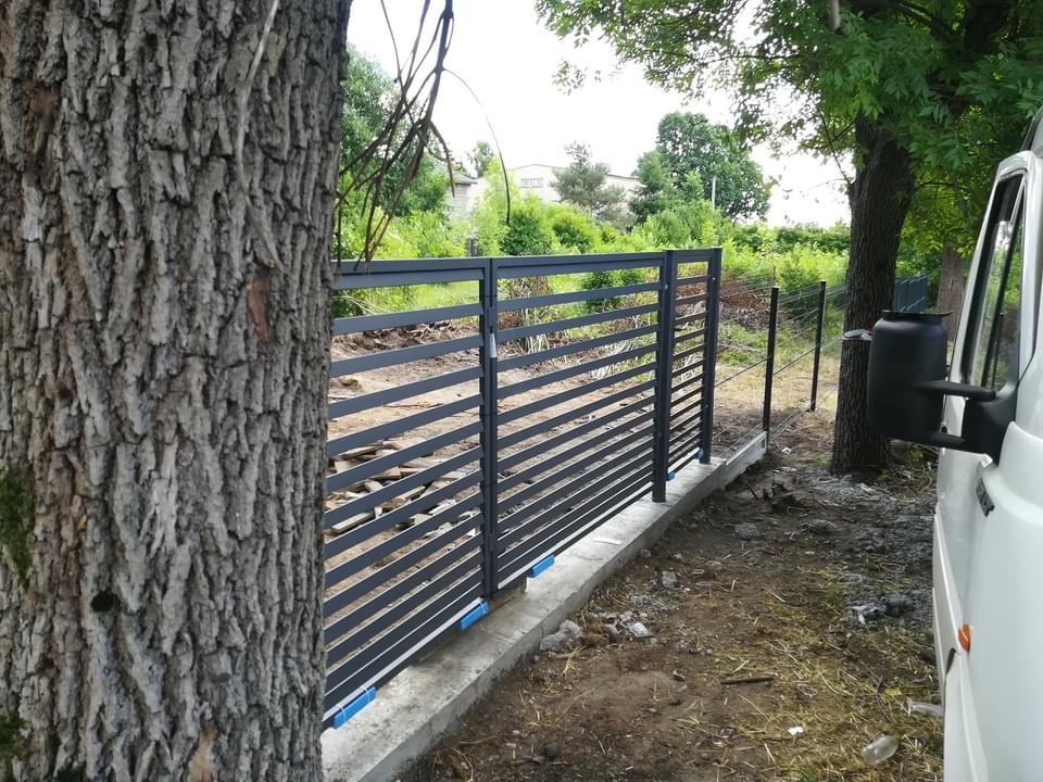 Nowoczesna, pozioma, metalowa brama w kolorze grafitowym, zamontowana na betonowej podmurówce z niebieskimi podkładkami, widoczna zza pnia drzewa, z samochodem dostawczym w tle.