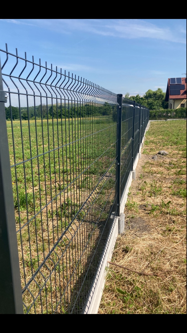 Nowoczesne ogrodzenie panelowe z podmurówką betonową, widok z perspektywy gruntu, na tle zielonego pola i domu z panelami fotowoltaicznymi na dachu.