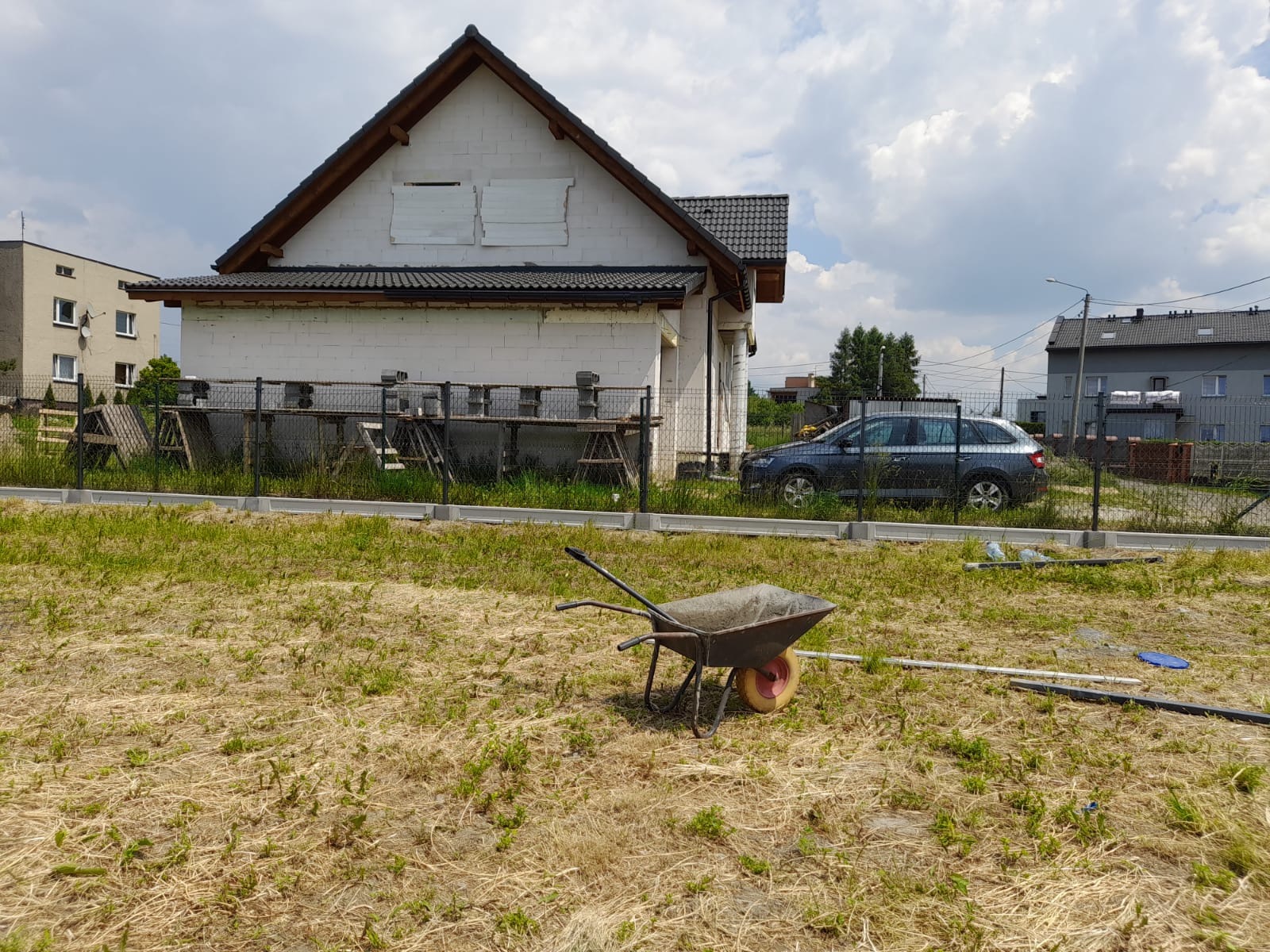 Dom w trakcie budowy z białego bloczka, widoczne rusztowania, taczka na pierwszym planie, teren częściowo porośnięty trawą, szara osobówka za ogrodzeniem.