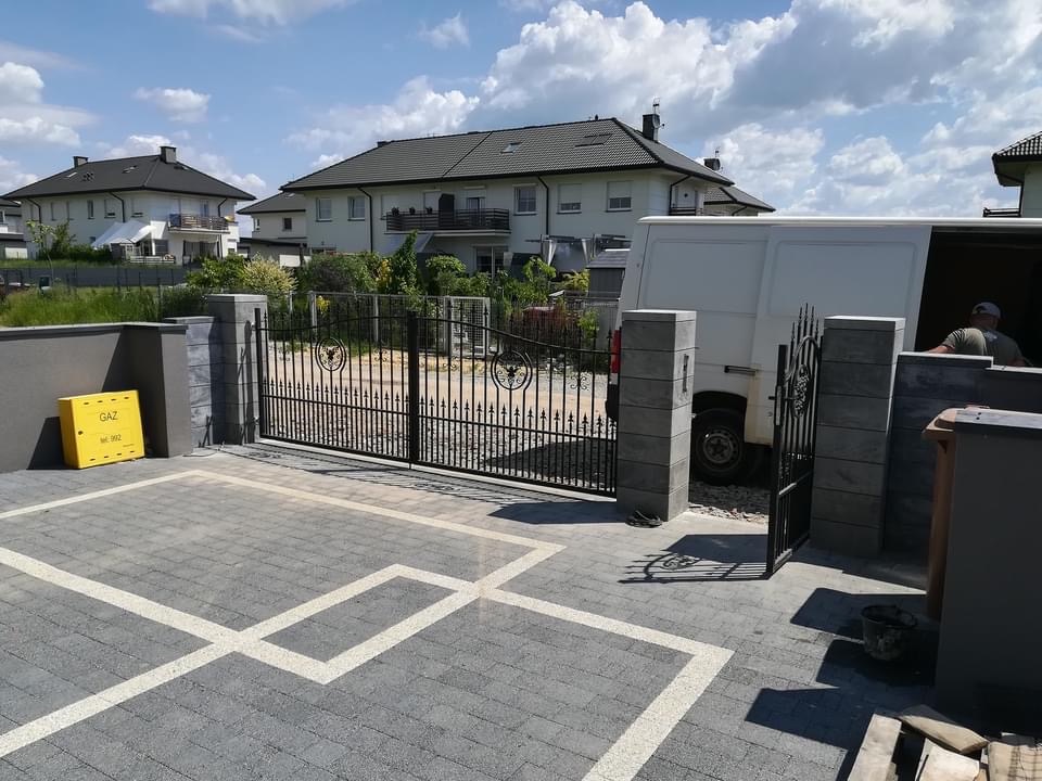 Czarna, metalowa brama wjazdowa z dekoracyjnymi elementami, zamontowana na posesji w Tarnowskich Górach, z widocznym fragmentem białego samochodu dostawczego w tle.