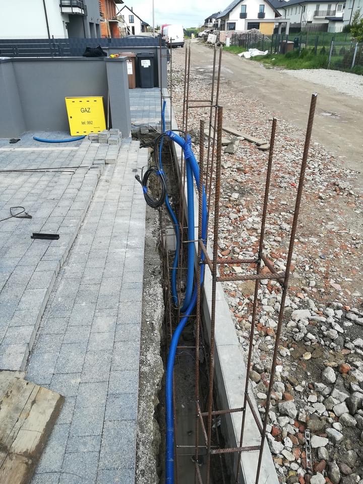 Przygotowanie fundamentów pod elewację z widocznym zbrojeniem, niebieskimi rurami instalacyjnymi i częściowo ułożoną kostką brukową, żółta skrzynka gazowa w tle.