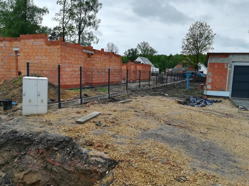 Budowa domu jednorodzinnego w trakcie realizacji: widoczne mury z czerwonej cegły, ogrodzenie z czarnej siatki oraz skrzynka elektryczna przyłączeniowa na placu budowy, w tle domy mieszkalne.