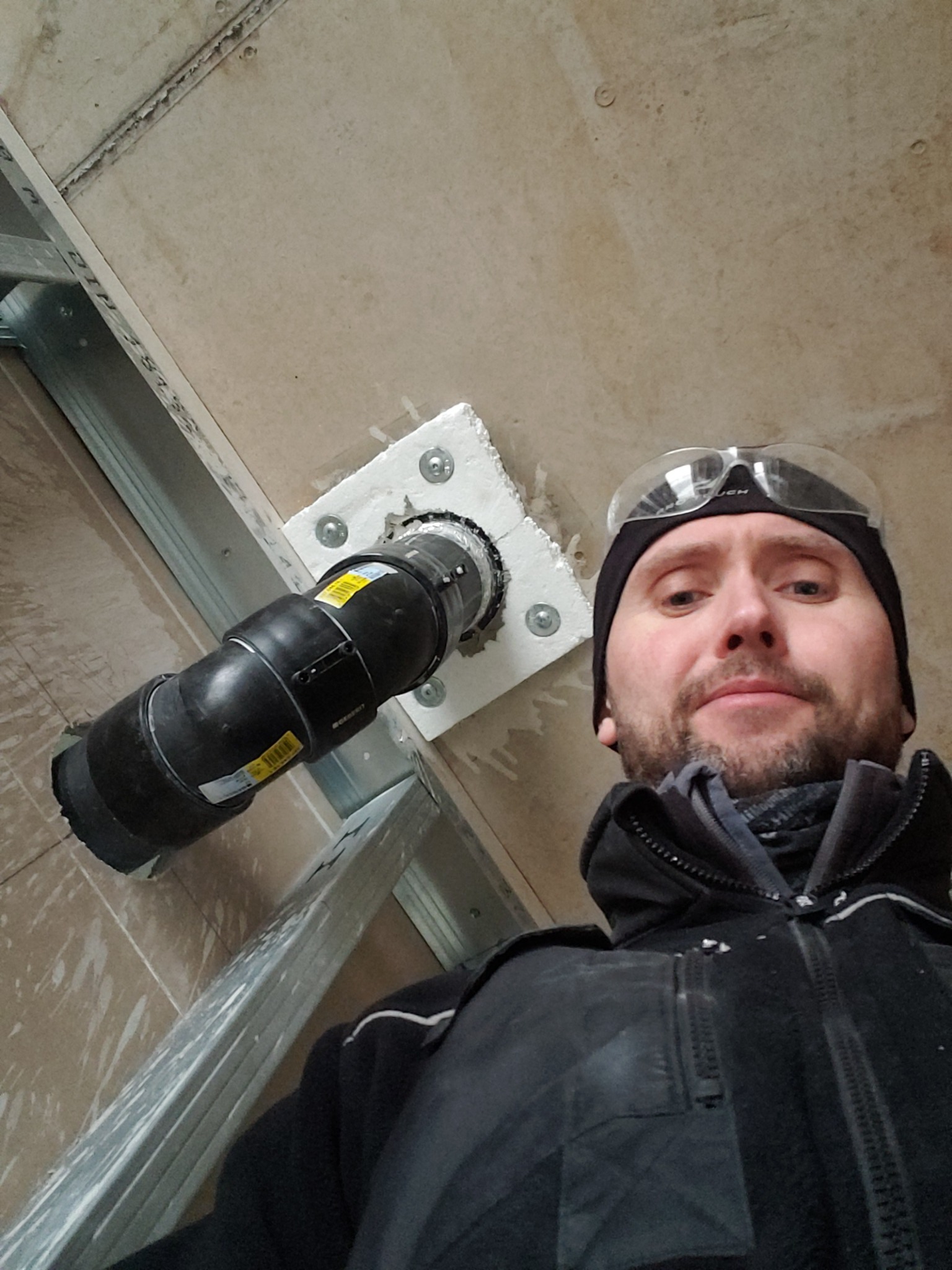 Hydraulik w okularach ochronnych, robiący selfie na tle zamontowanej czarnej rury kanalizacyjnej przechodzącej przez płytę gipsową w konstrukcji metalowej