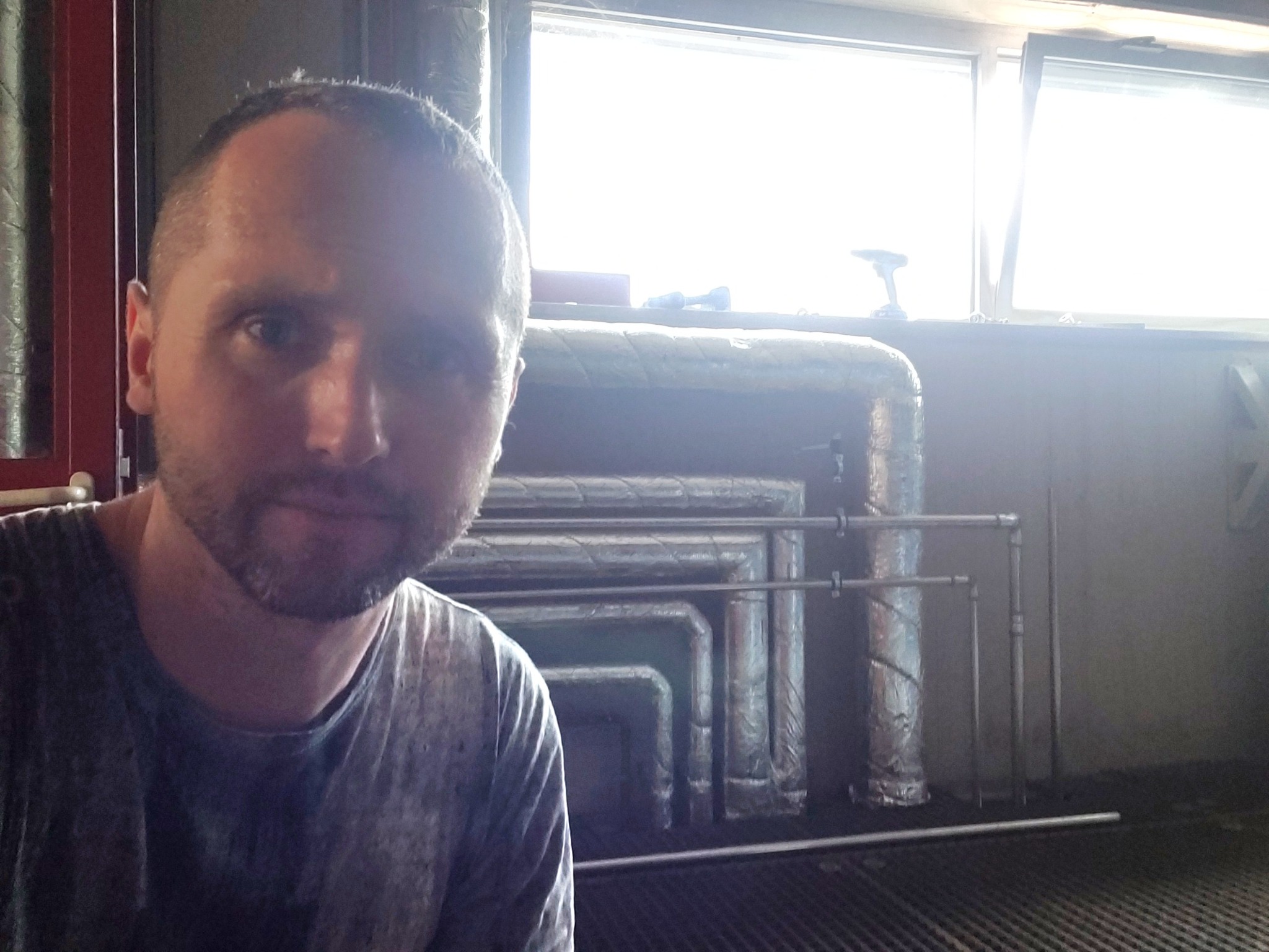 Mężczyzna pozuje na tle rur izolowanych folią aluminiową, widocznych w jasnym pomieszczeniu z oknem, na parapecie leży wiertarka.