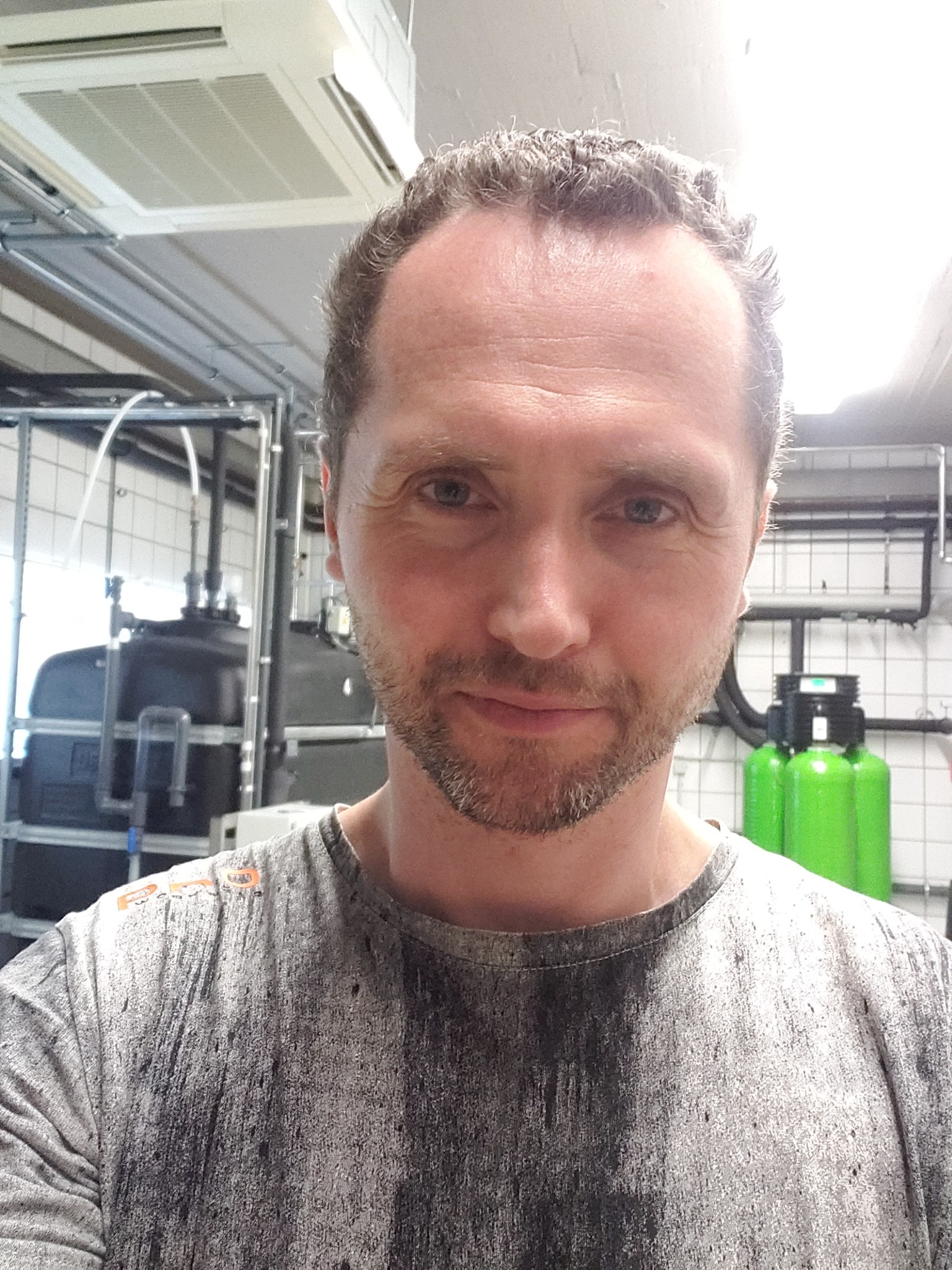 Mężczyzna robiący selfie na tle instalacji wodnej z czarnym zbiornikiem i zielonymi filtrami, widoczna klimatyzacja sufitowa.