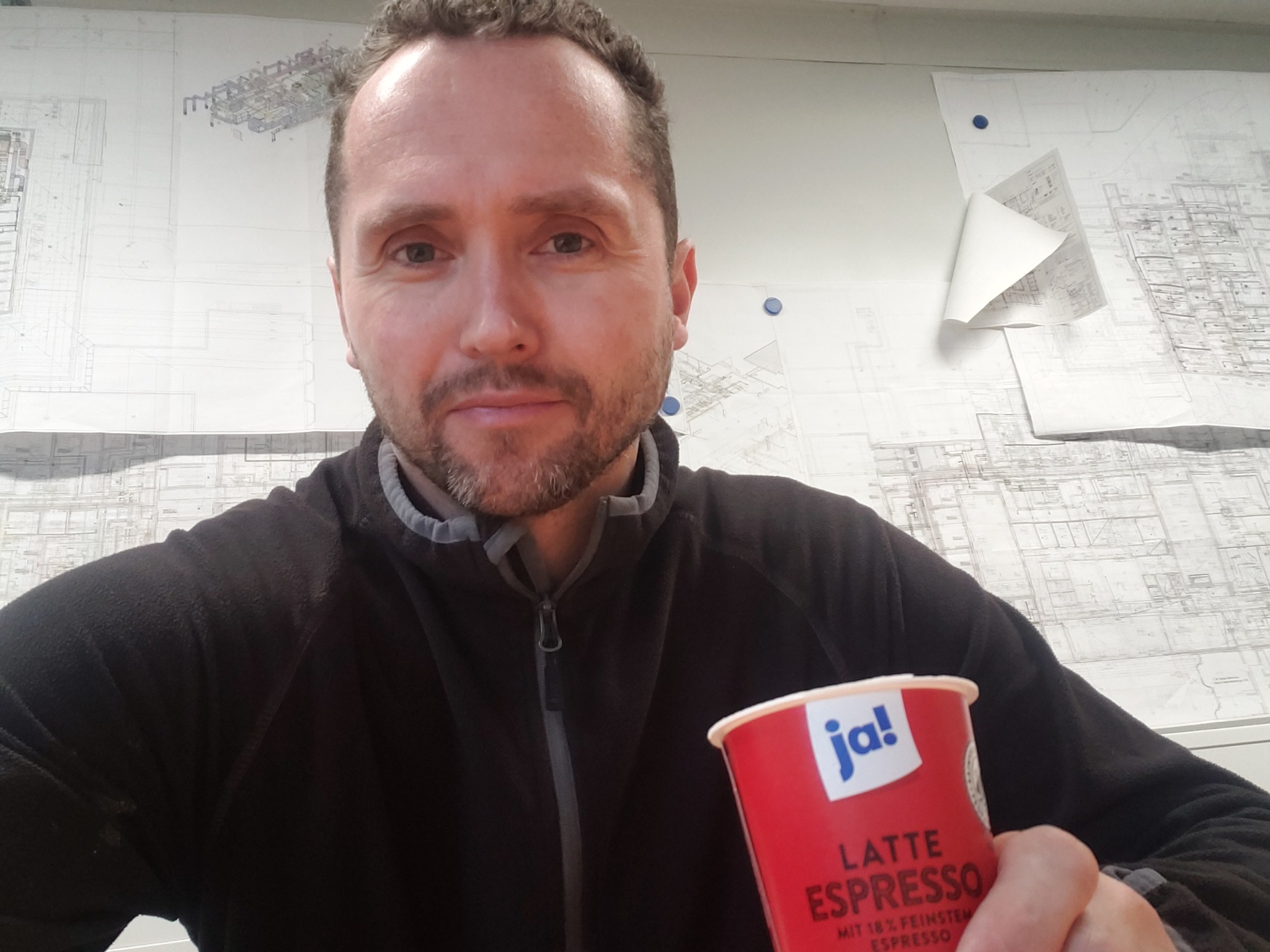 Mężczyzna z kawą na tle planów instalacji budowlanych przypiętych do ściany.
