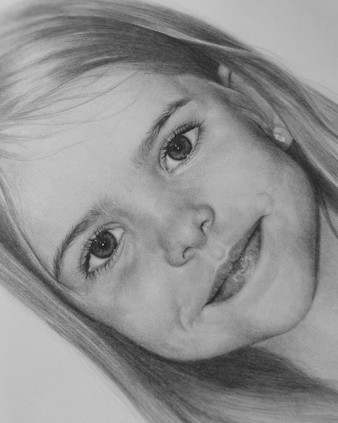 Czarno-biały portret ołówkiem dziewczynki z długimi włosami, uśmiechająca się, z delikatnym cieniowaniem i widoczną fakturą papieru.