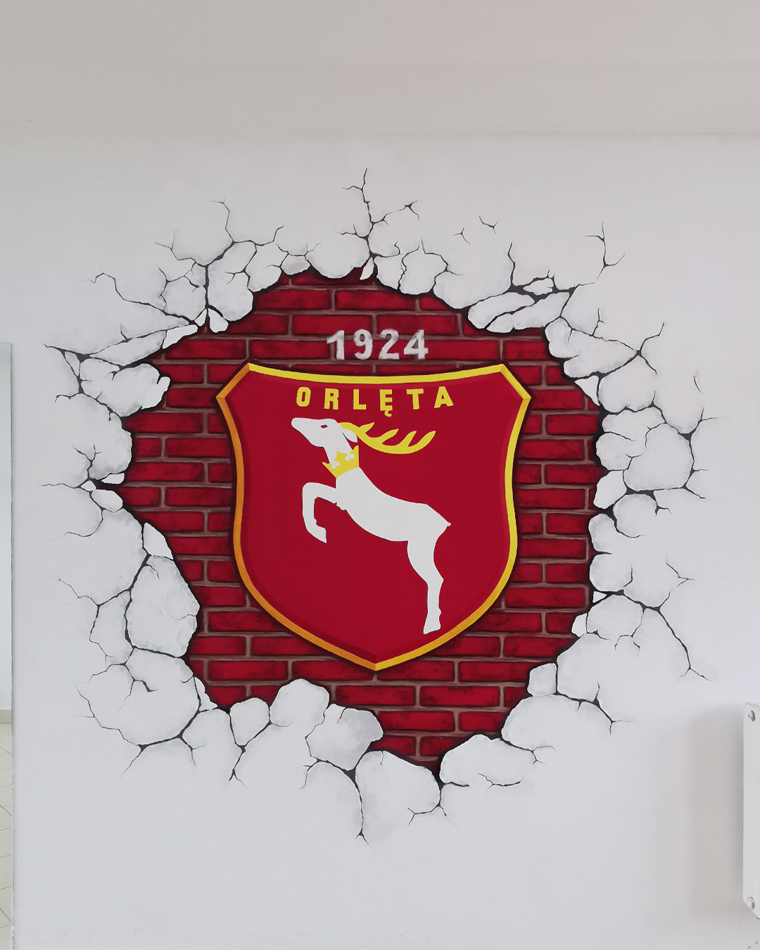 Artystyczne malowanie ściany przedstawiające herb 'Orlęta' z datą 1924, umieszczony na tle imitującym czerwoną cegłę, z efektem pęknięć na białej ścianie.