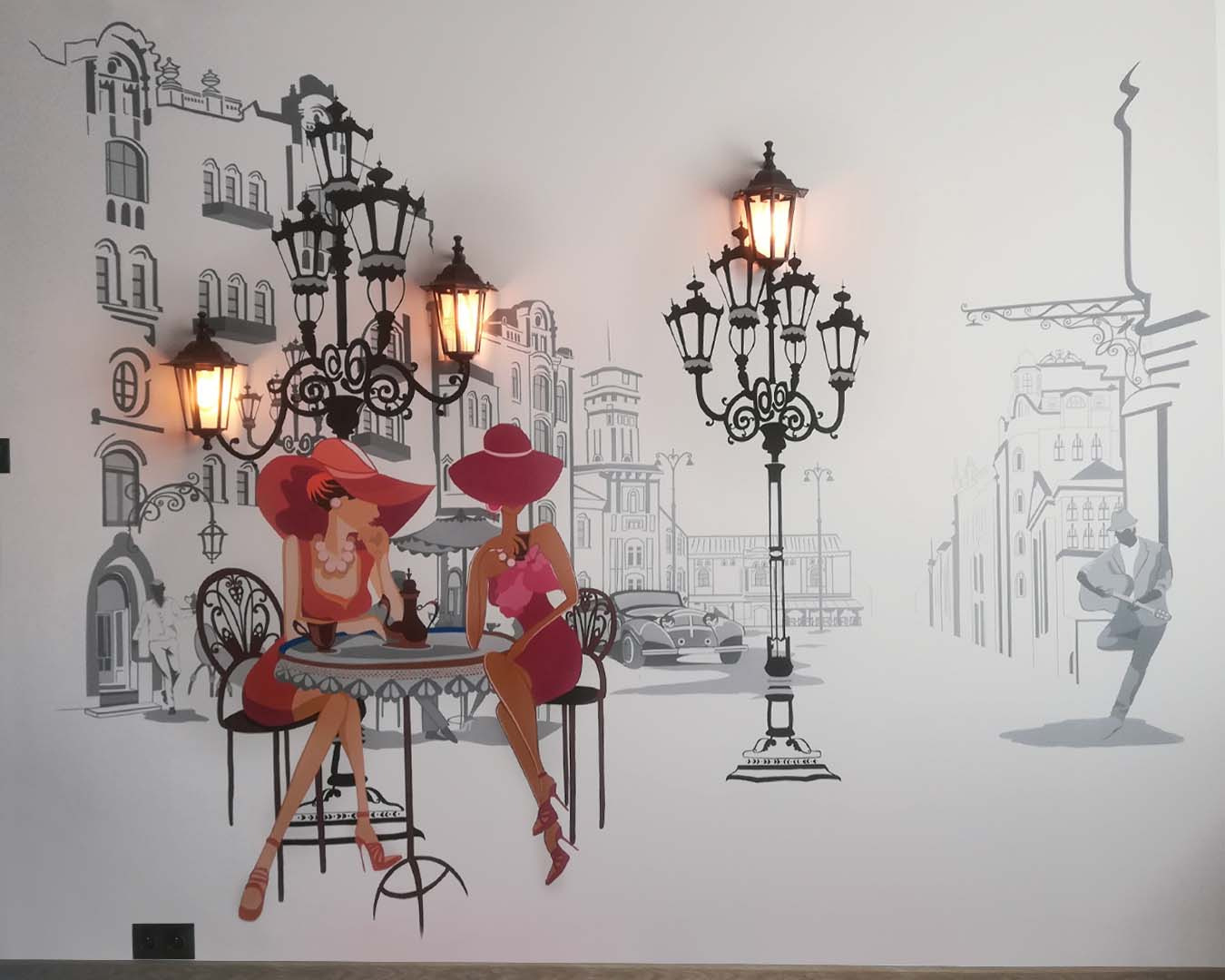 Artystyczne malowanie ściany przedstawiające dwie kobiety w kapeluszach siedzące przy stoliku w kawiarni w stylu retro, z elementami architektury miejskiej i latarniami ulicznymi w tle.