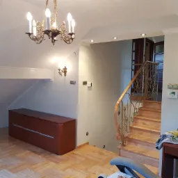 Jasne wnętrze domu w trakcie wykańczania: drewniane schody z ozdobną balustradą, parkiet w jodełkę, komoda w kolorze ciemnego drewna i żyrandol w stylu retro.