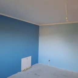 Świeżo pomalowane pomieszczenie w odcieniach błękitu, z widoczną puszką instalacyjną na ścianie i surową podłogą w trakcie wykończenia.