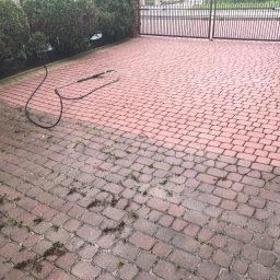 Paint Services Kamil Iwosa - Częściowo umyta, czerwona kostka brukowa z zielonymi obrzeżami, z widocznym wężem ciśnieniowym i zanieczyszczeniami, prowadząca do metalowej bramy.