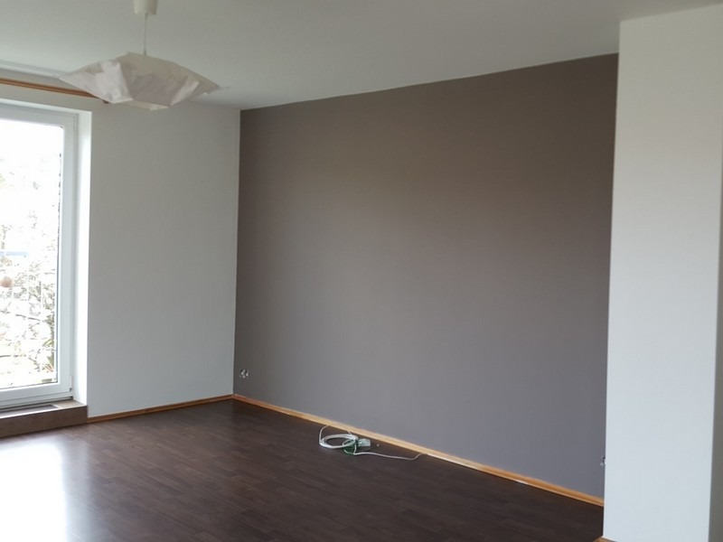 Pomieszczenie po malowaniu: brązowa ściana kontrastuje z bielą, ciemna podłoga, okno z widokiem na zieleń. Minimalistyczna lampa dodaje nowoczesności.