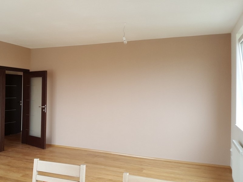 Jasny pokój po malowaniu ścian na beżowo, z drewnianą podłogą i drzwiami z przeszkleniami. Widoczny fragment krzesła i grzejnik pod oknem.