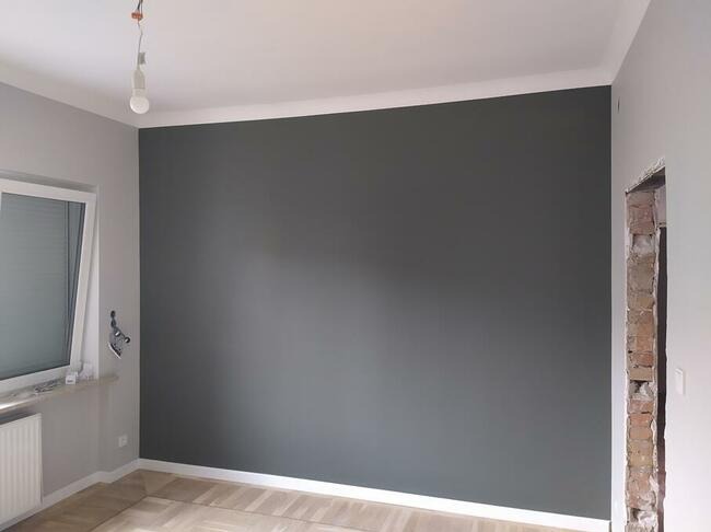 Pomieszczenie po malowaniu: szara ściana z białą listwą, parkiet, okno z żaluzjami i fragment ściany z odsłoniętą cegłą. Widok na odnowione wnętrze.