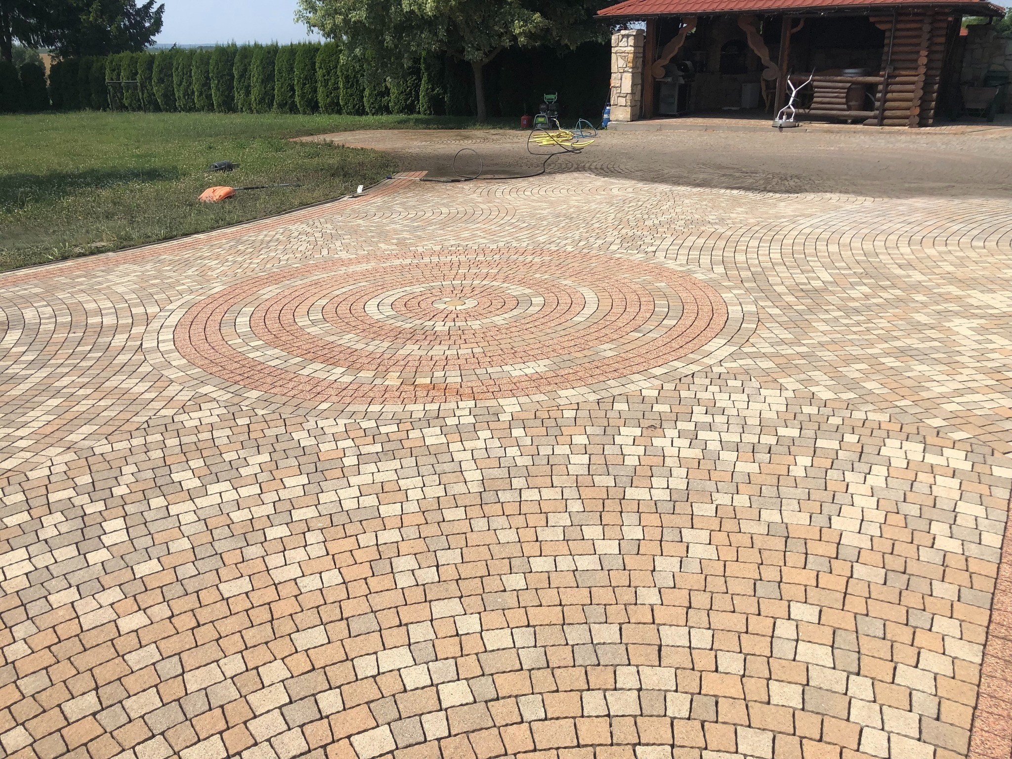 Wzorzysta kostka brukowa ułożona w koncentryczne okręgi, otoczona zielenią i elementami architektury ogrodowej. Widoczny fragment węża i myjki ciśnieniowej.