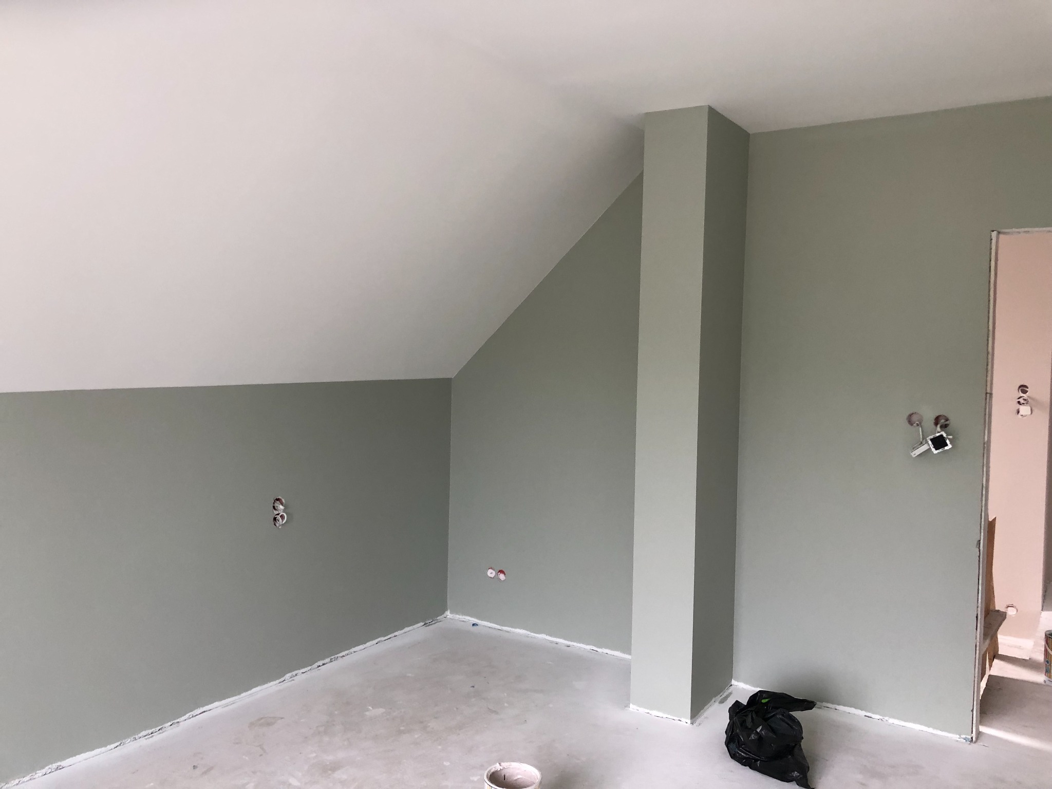 Pomieszczenie po malowaniu ścian na odcień zieleni, widoczne gniazdka elektryczne, sufit biały, podłoga przygotowana do dalszych prac wykończeniowych.