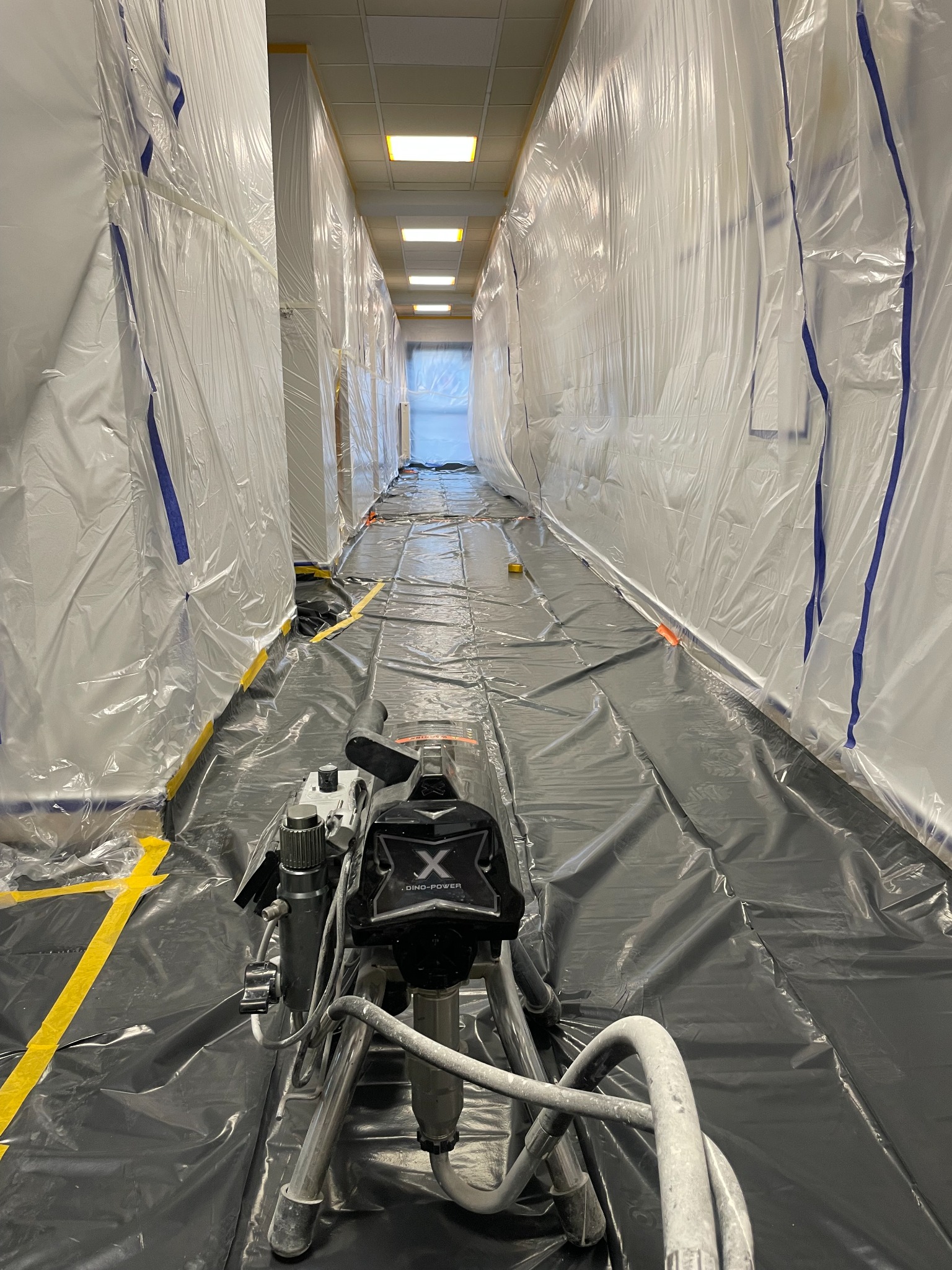 Korytarz biurowy przygotowany do malowania natryskowego, zabezpieczony folią i taśmą. Widoczny agregat malarski Dino-Power na czarnej folii ochronnej.