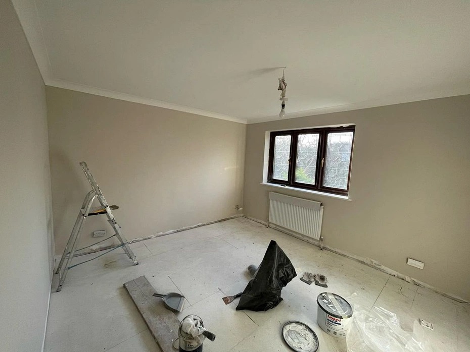 Pokój po malowaniu ścian na beżowo, widoczna drabina, pędzle, wiadro z farbą i okno. Ściany świeżo pomalowane, zabezpieczona podłoga folią malarską.